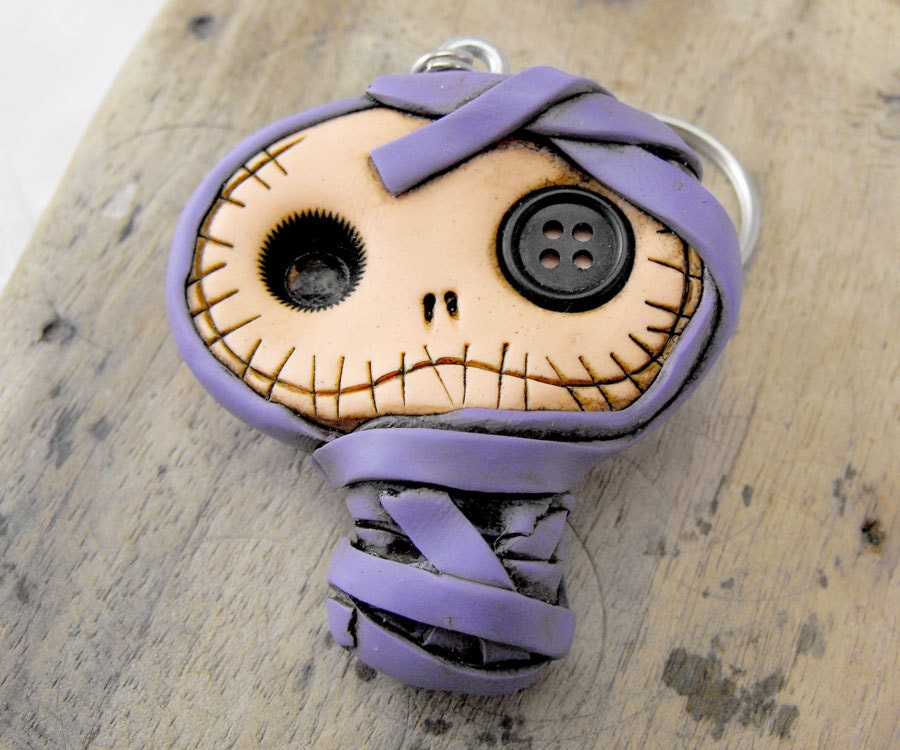 Sweet baby mummy keychain in dirty purple. Creepy and cute baby skull with a pink button inside his eye.