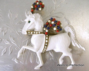 Beautiful unique vintage horse brooch signed Jeanne. White horse with red, white and blue cabochon accents. Gold tone brooch painted white. Beautifully done and signed Jeanne. Measures 2-1/2 by 2-5/8 inches. Secure safety clasp. In very good condition. Little is known about the Jeanne company. It's figural jewelry is highly collectible and always beautifully made.