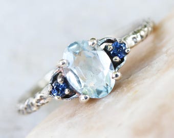 Oval Blue topaz ring with blue sapphire side set gems in prongs setting on sterling silver oxidized hard textured band